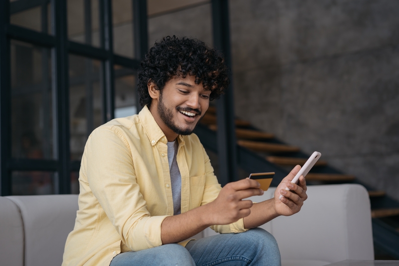 a man sitting on a couch with a credit card in one hand a cell phone in the other wondering how long a credit card balance transfer takes