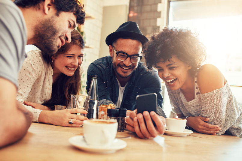 Portrait of cheerful young friends looking at smartphone while sitting in a cafe. Mixed race people sitting at a table in a restaurant using mobile phone.