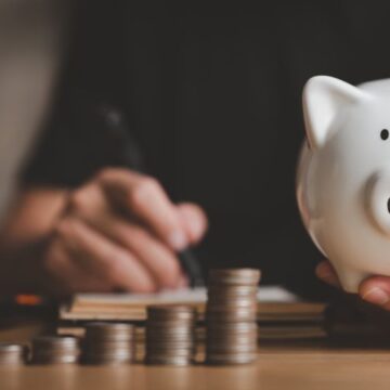 Person holding a money bank with a stack of coins in the background